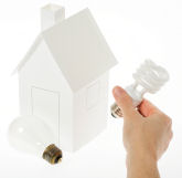 Photo of small white house with incandescent light bulb along with a person's hand holding a compact fluorescent light bulb.