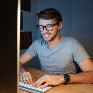 Man with glasses sits in front of a computer, smiling