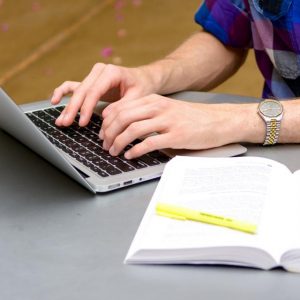 Hands type at a laptop, with a notebook and highlighter in the foreground
